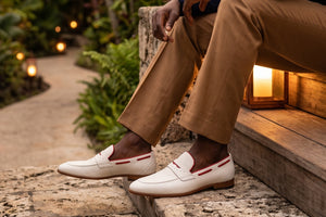A person seated on outdoor stone steps wearing tan trousers and white penny loafers with thin red stripes, photographed in a warm garden setting with lanterns and blurred greenery in the background.