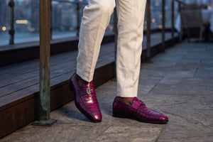 A person wearing bright magenta crocodile-embossed loafers with a gold-tone logo on the side, paired with off-white trousers, standing on a stone deck next to a wooden railing at dusk.