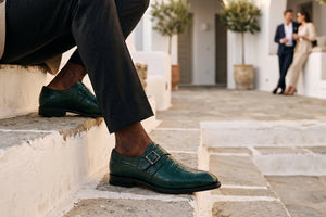 A close-up lifestyle shot of a man wearing dark green crocodile-embossed double strap loafers with black trousers, sitting on white stone steps in a Mediterranean-style courtyard.