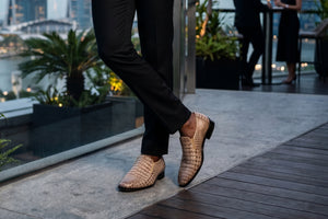 A person wearing black trousers and tan crocodile-embossed slip-on loafers on a rooftop terrace with a city skyline in the background.