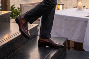 A person wearing dark charcoal trousers and dark burgundy crocodile-embossed leather loafers, sitting on dark stone steps next to a table with a white tablecloth and a lit candle.