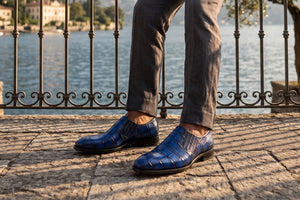 A person wearing blue crocodile-embossed leather slip-on shoes and gray trousers, standing on a stone terrace with a lake and wrought-iron railing in the background.