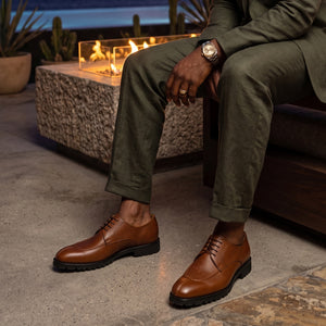 Person wearing brown leather shoes and green pants sitting by a fire pit.