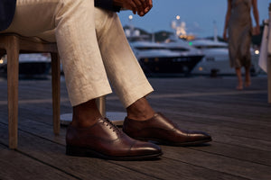 A close-up lifestyle shot of a man wearing brown leather cap-toe oxford shoes and cream linen trousers, seated on a wooden chair on a wooden dock with a marina and yachts in the background at dusk.