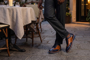 A man wearing dark gray trousers and distinctive brown and navy wingtip oxford shoes, standing on a stone patio next to a dining table at an outdoor restaurant.